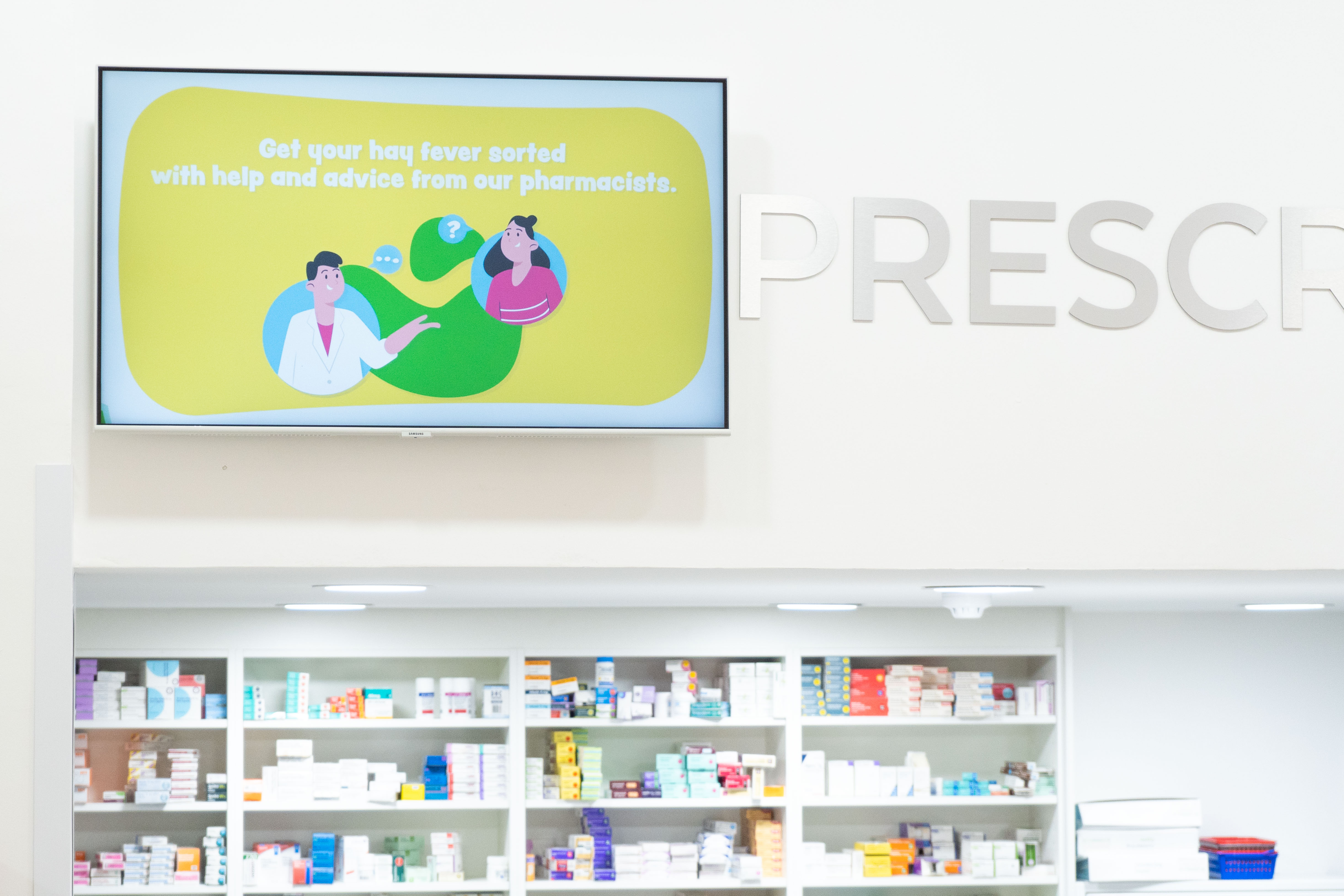 Top section of pharmacy front desk, showing TV at top of wall with graphic image on, and top of pharmacy medication shelves