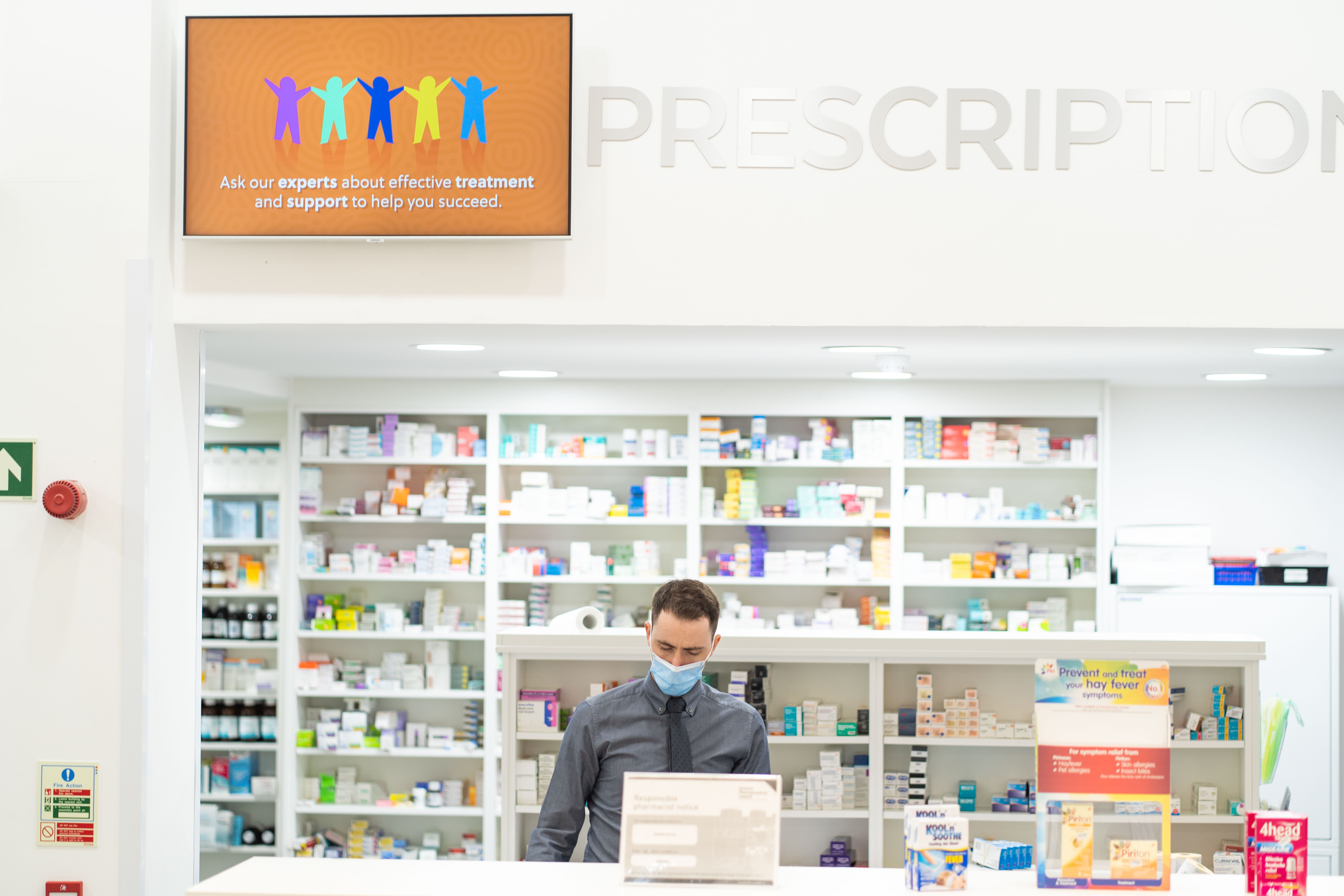 Male pharmacist wearing mask, looking down at till, medication shelves behind him and TV above him showing graphic image