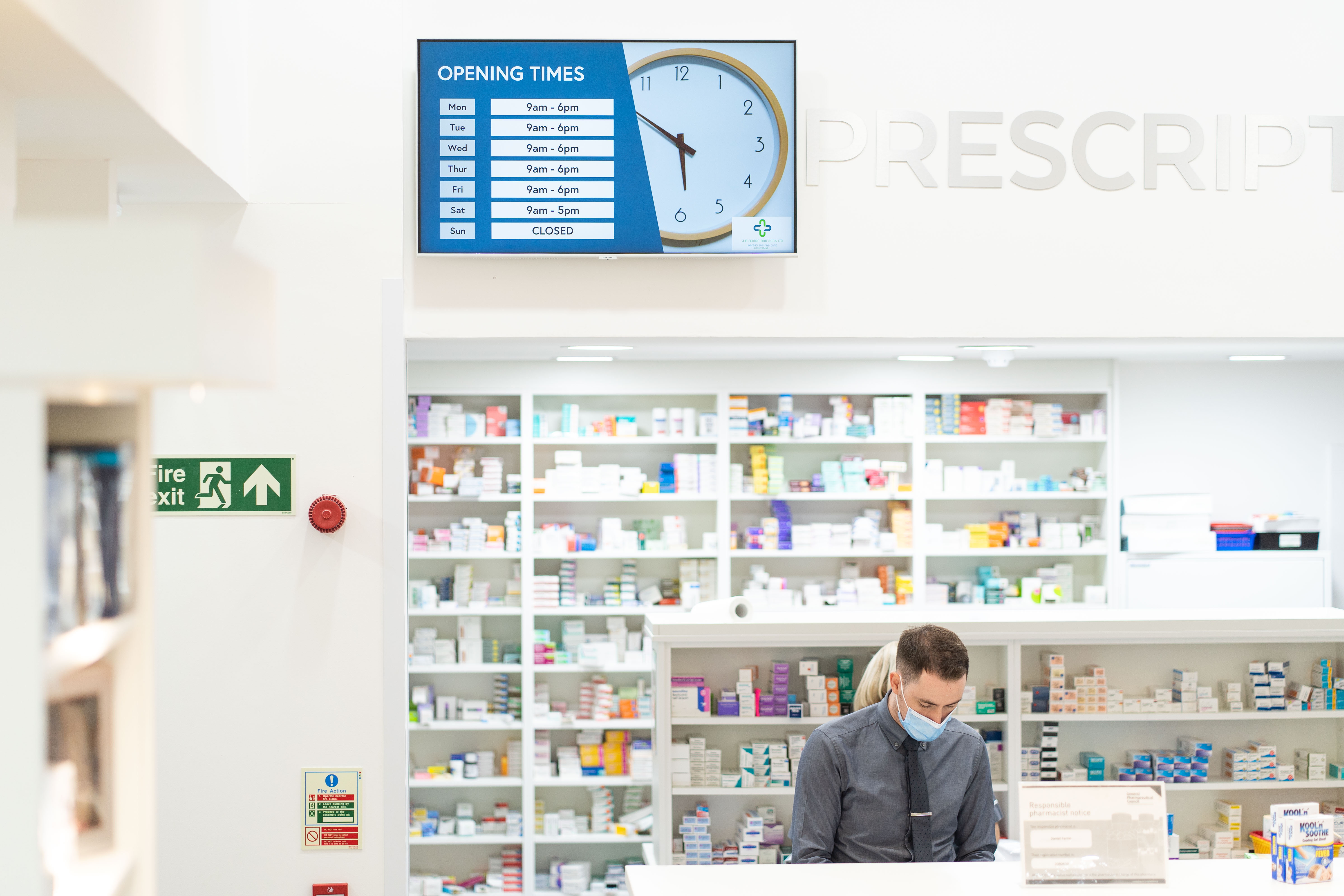 Image of male pharmacist wearing a mask, stood at till in pharmacy with medication shelves behind him