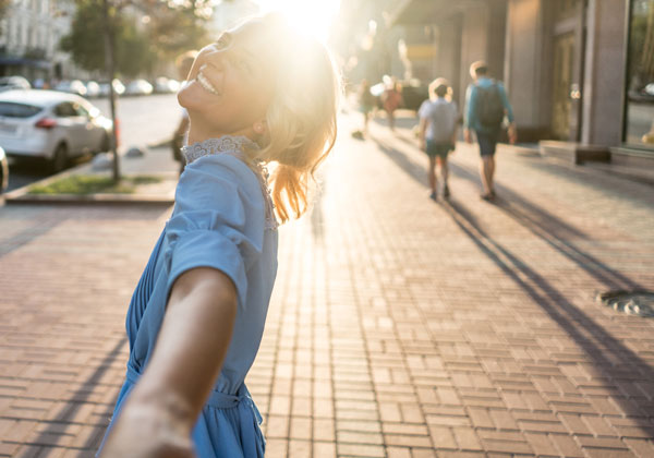 Smiling woman in blue dress stood on street side on, arm stretched toward camera and sun shining behind her