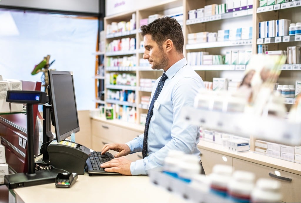 Man stood at pharmacy front desk with both hands on keyboard, looking at screen