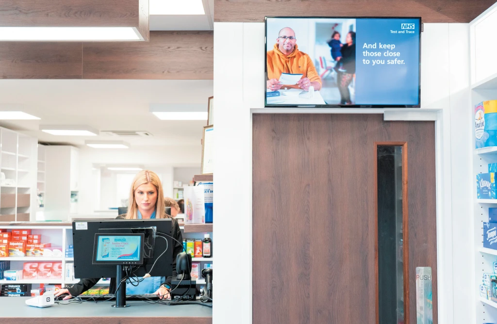 pharmacy display screen and female pharmacist stood at till